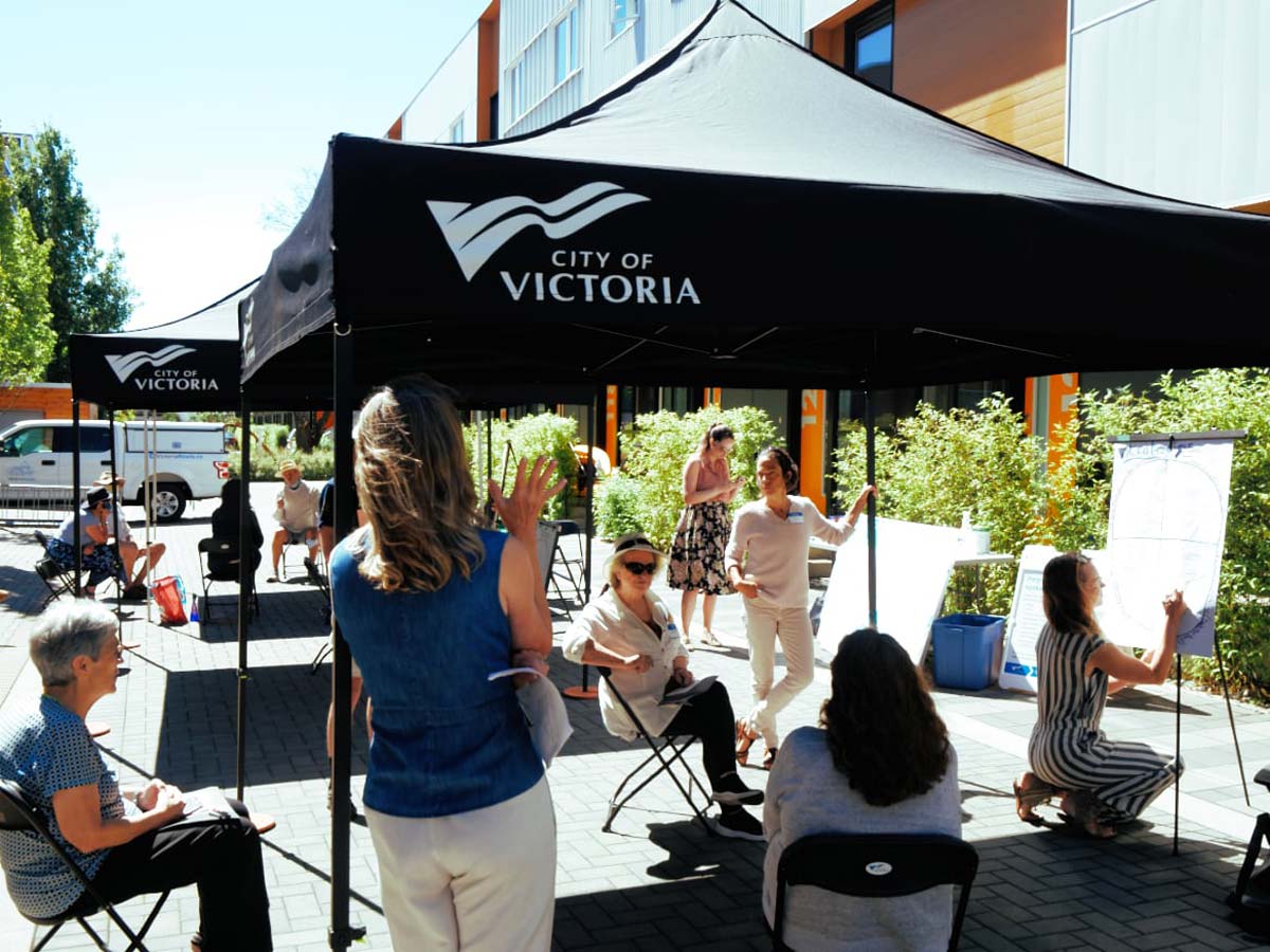 Outdoor large shade tent with the City of Victoria logo with a bunch of people sitting under it and around it.
