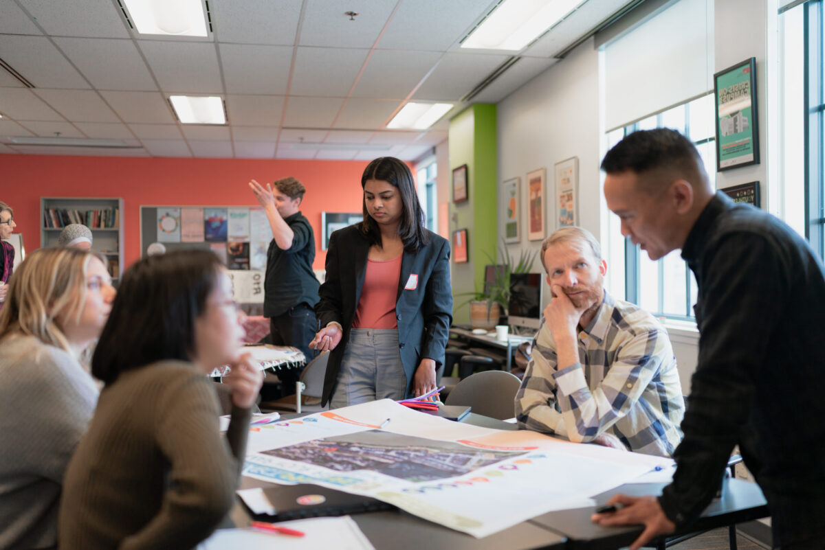 Facilitator stands up in a buzzing room with people standing around tables collaborating.