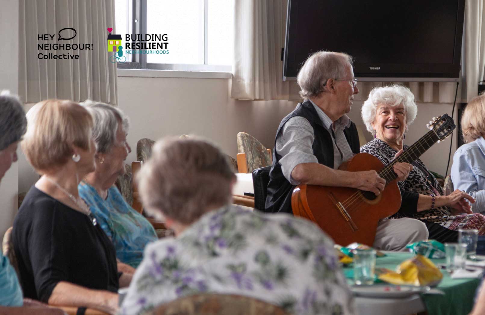 A neighbour plays guitar at the table with a bunch of old folks.