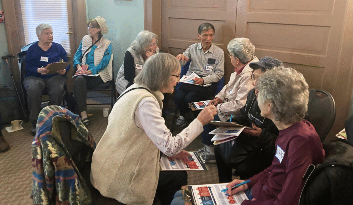 A diverse group of eight people sits on folding chairs in a room with beige doors and light green walls. They are talking animatedly with one another.