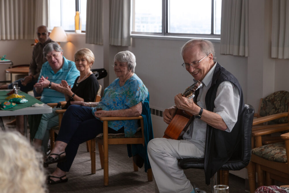 An elderly pale-skinned man plays guitar in a chair at the front of a room with a long table. Three other elders are sitting at the table and are clapping and smiling.