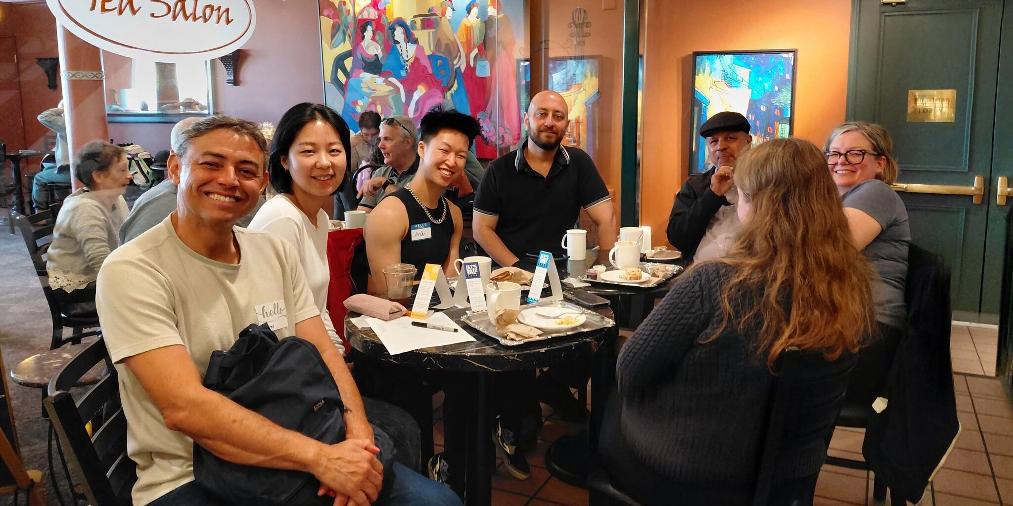 Group of seven people sitting around a table at a cafe looking at and smiling at the camera.