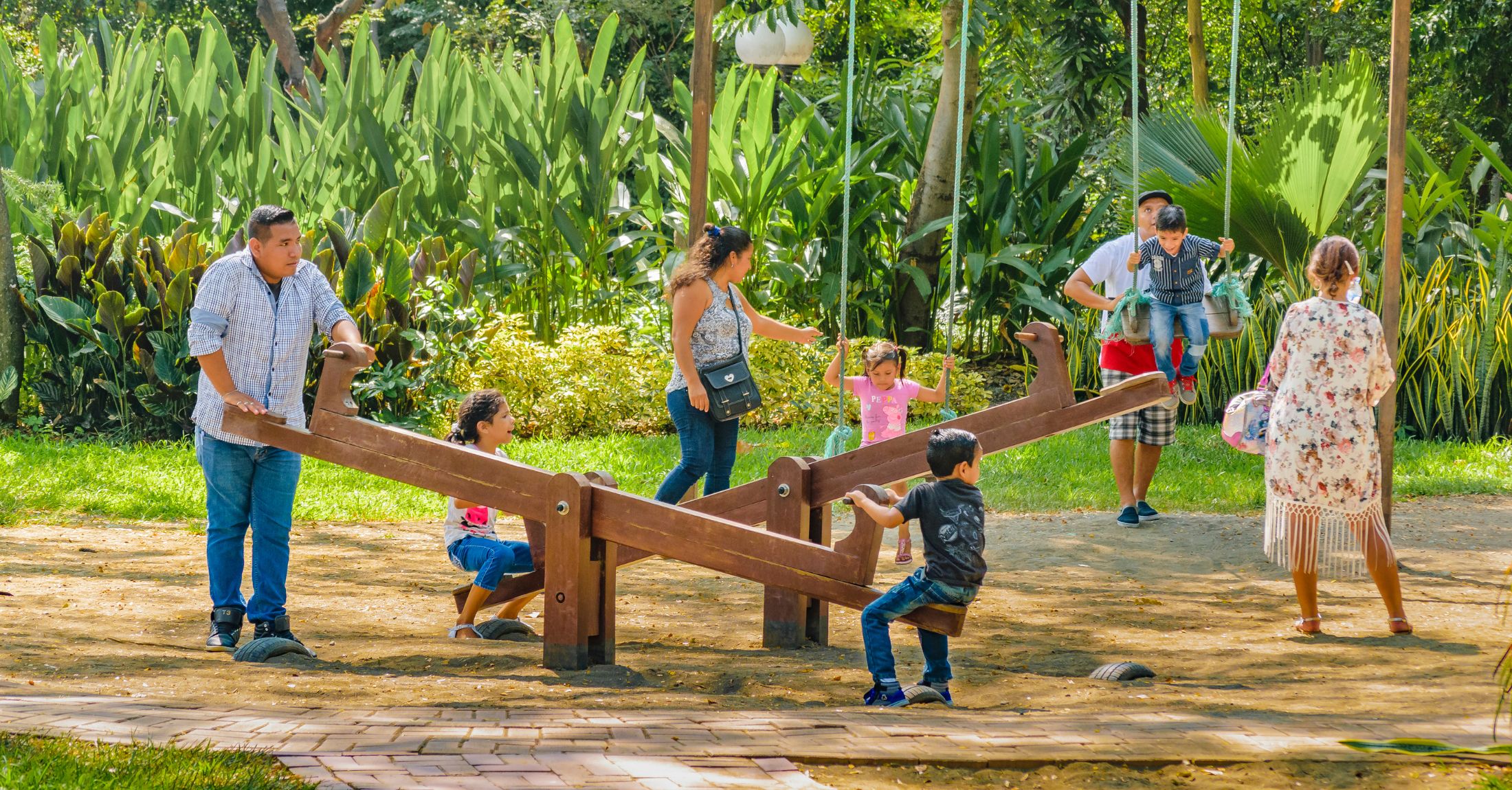 Parents playing with their children at a playground with seesaws and swings.